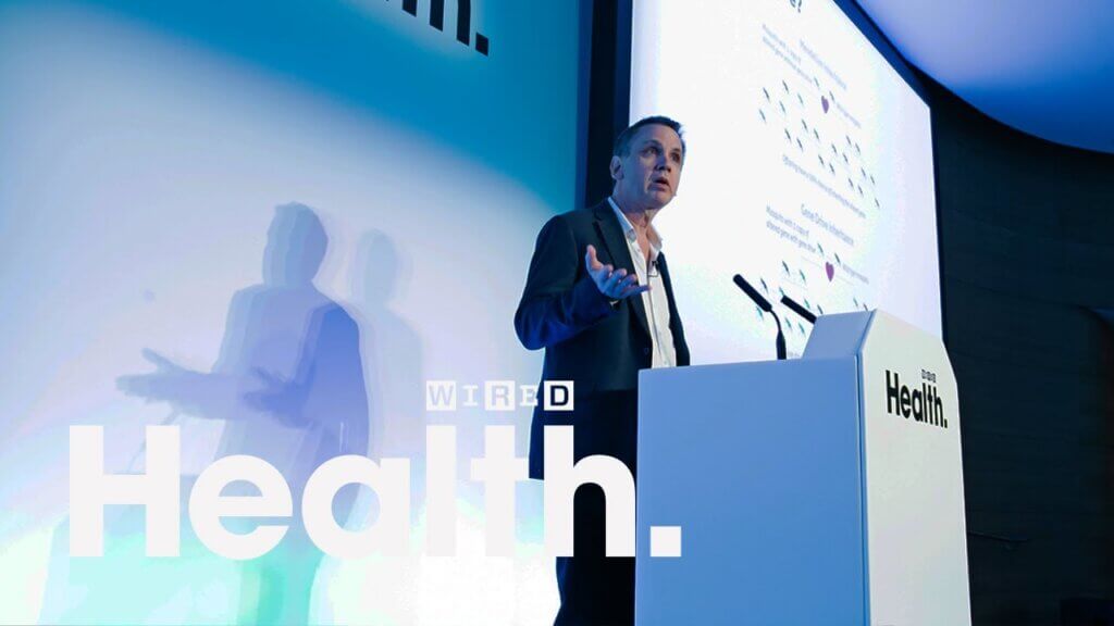 A man speaks at a podium with a Wired Health logo on it. Behind him is a large screen displaying a presentation, and the words "Health." and "WIRED" are prominently visible. The setting appears to be a professional conference or event dedicated to health topics.