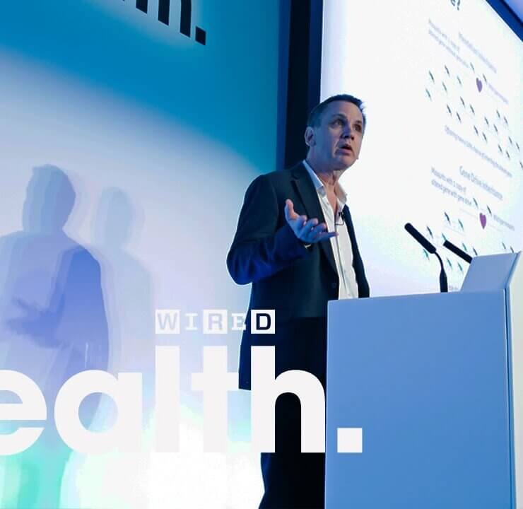 A man speaks at a podium with a Wired Health logo on it. Behind him is a large screen displaying a presentation, and the words "Health." and "WIRED" are prominently visible. The setting appears to be a professional conference or event dedicated to health topics.