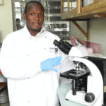 Charles Batume standing beside a microscope in a lab