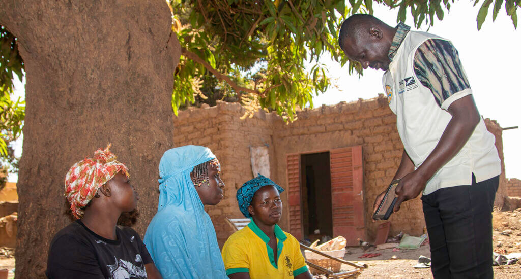 In Burkina Faso, A Target Malaria team member shows 3 women information on an a tablet. The women are sitting under a tree in front o a brick building.